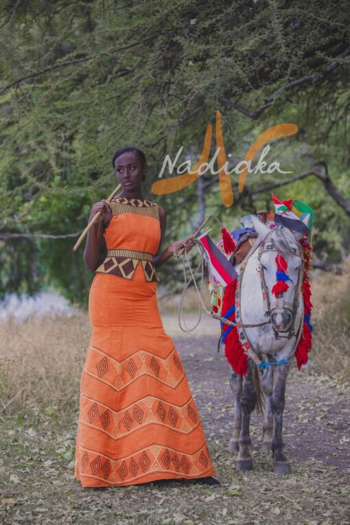Robe longue brodée, en pagne tissé orange à brillantine or, Avec ceinture large brodée. Foulard et chapeau brodé assortis.