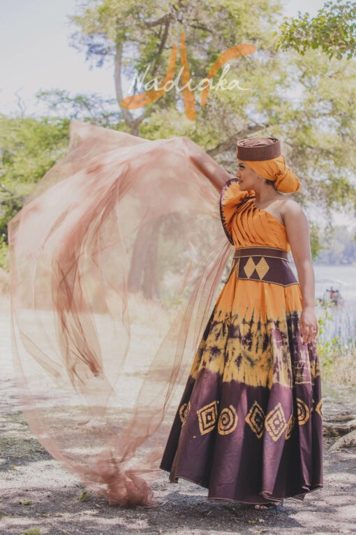 Robe longue, haut drapé, en tissu batik couleur moutarde et marron. Avec ceinture large brodée et tulles traîne brillant marron. Foulard et chapeau brodé assortis.