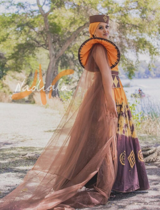 Robe longue, haut drapé, en tissu batik couleur moutarde et marron. Avec ceinture large brodée et tulles traîne brillant marron. Foulard et chapeau brodé assortis.