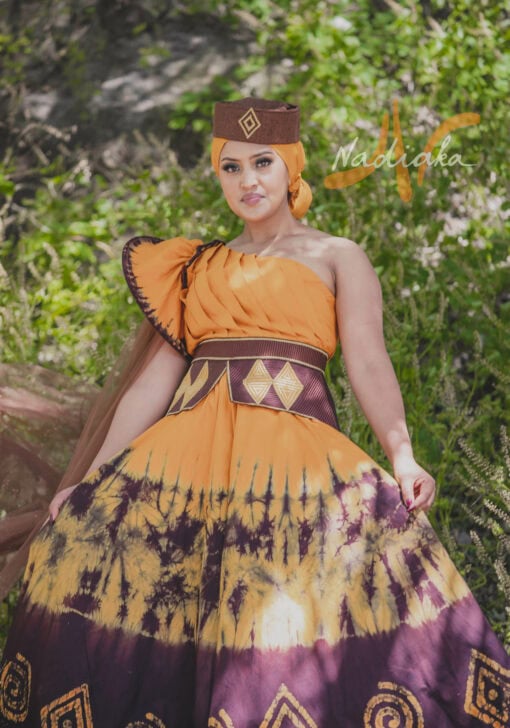 Robe longue, haut drapé, en tissu batik couleur moutarde et marron. Avec ceinture large brodée et tulles traîne brillant marron. Foulard et chapeau brodé assortis.