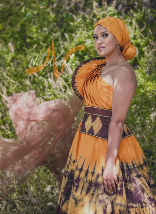 Robe longue, haut drapé, en tissu batik couleur moutarde et marron. Avec ceinture large brodée et tulles traîne brillant marron. Foulard et chapeau brodé assortis.