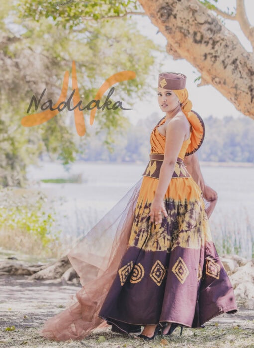 Robe longue, haut drapé, en tissu batik couleur moutarde et marron. Avec ceinture large brodée et tulles traîne brillant marron. Foulard et chapeau brodé assortis.