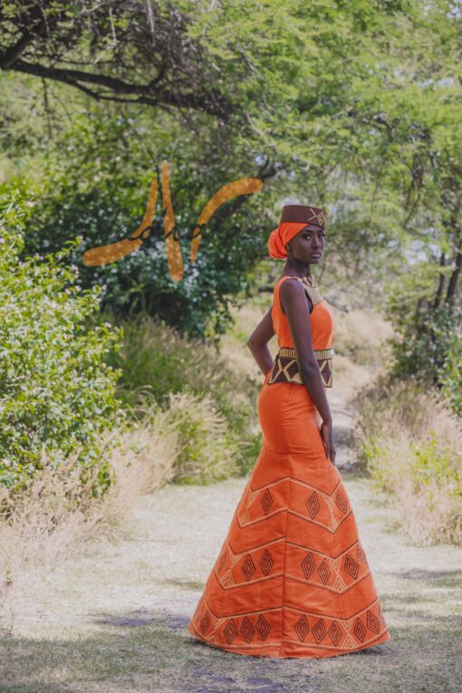 Robe longue brodée, en pagne tissé orange à brillantine or, Avec ceinture large brodée. Foulard et chapeau brodé assortis.