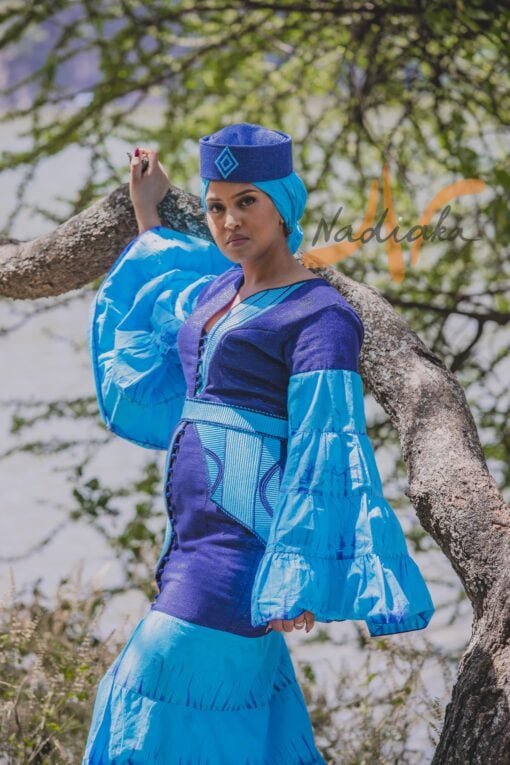 Robe longue volantée, brodée, en pagne tissé à brillantine, mix tissu batik couleur bleue turquoise et bleu. Avec ceinture large asymétrique brodée. Foulard et chapeau brodé assortis.