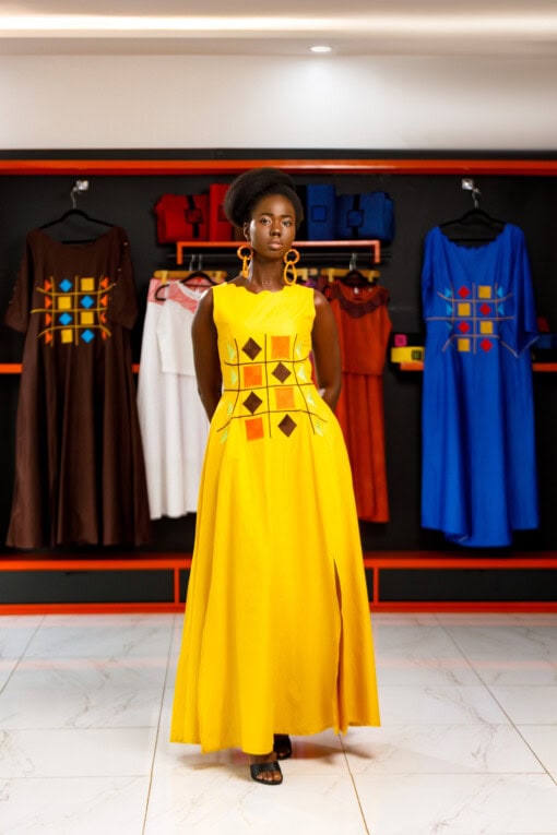 Longue robe brodé de couleur jaune moutarde sans manches en tissu coton woodin avec des broderies à l'avant. La robe est agrémentée de fentes en bas sur chaque coté.