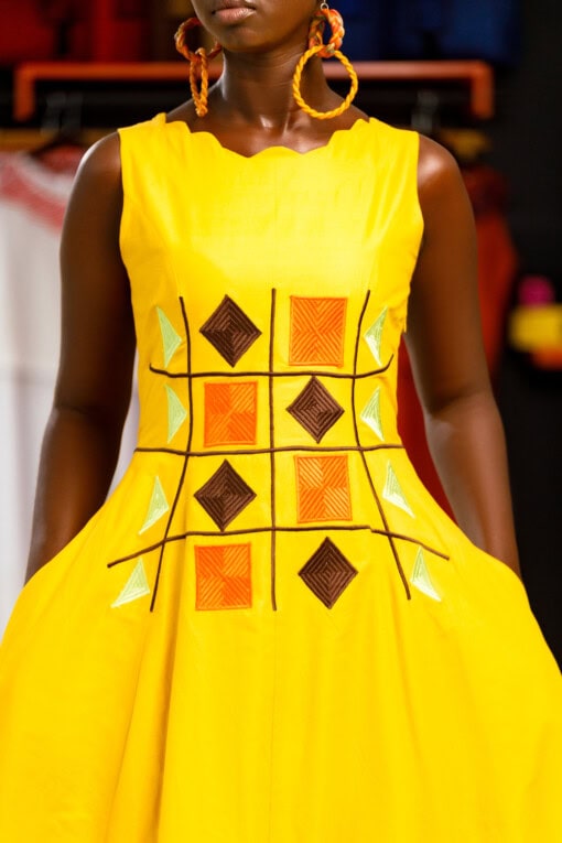Longue robe brodé de couleur jaune moutarde sans manches en tissu coton woodin avec des broderies à l'avant. La robe est agrémentée de fentes en bas sur chaque coté.